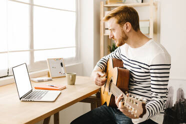 Man learning guitar through online tutorial on laptop sitting at table - XLGF02876