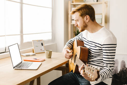 Mann lernt Gitarre mit Hilfe eines Online-Tutorials am Laptop am Tisch sitzend - XLGF02876