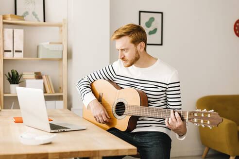 Junger Mann mit Gitarre E-Learning über Laptop am Tisch sitzend - XLGF02877