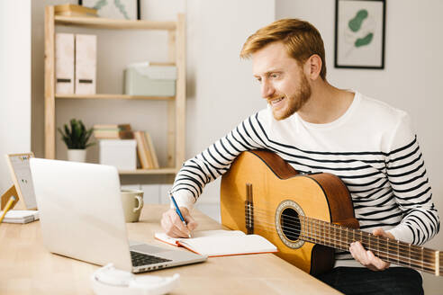 Lächelnder Mann mit Gitarre, der am Tisch sitzend eine Musiknote in einen Laptop schreibt - XLGF02879