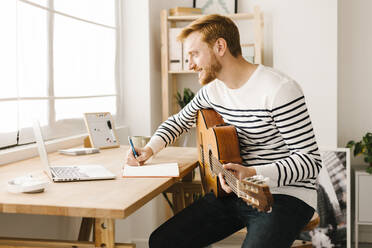 Lächelnder junger Mann mit Gitarre, der Musiknoten schreibt und zu Hause am Laptop lernt - XLGF02880