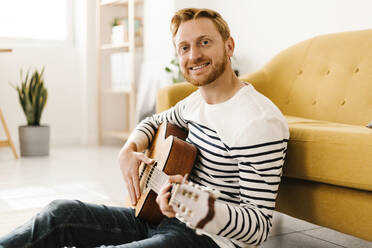 Lächelnder junger Mann mit Gitarre auf dem Sofa im Wohnzimmer - XLGF02885