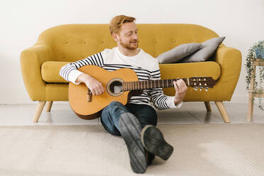 Junger Mann spielt Gitarre und sitzt zu Hause vor dem Sofa - XLGF02886