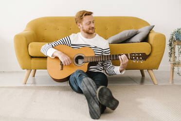 Young man playing guitar sitting in front of sofa at home - XLGF02886