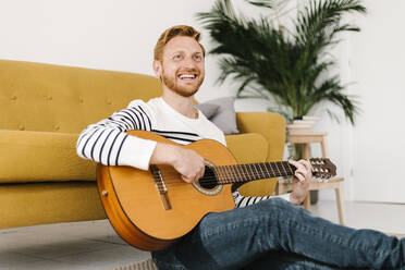 Cheerful man with guitar sitting in front of sofa at home - XLGF02887