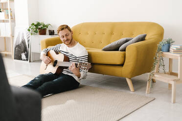 Junger Mann mit braunen Haaren übt Gitarre und sitzt zu Hause vor dem Sofa - XLGF02889