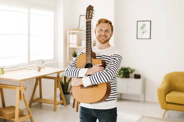 Glücklicher junger Mann mit Gitarre im Wohnzimmer zu Hause stehend - XLGF02890