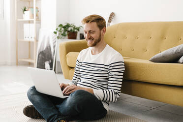 Lächelnder junger Mann mit Laptop im Schneidersitz vor dem Sofa zu Hause - XLGF02891