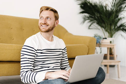 Glücklicher junger Mann mit Laptop im heimischen Wohnzimmer sitzend - XLGF02894