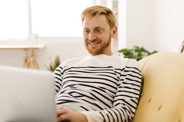 Lächelnder junger Mann mit Laptop auf dem Sofa zu Hause - XLGF02895