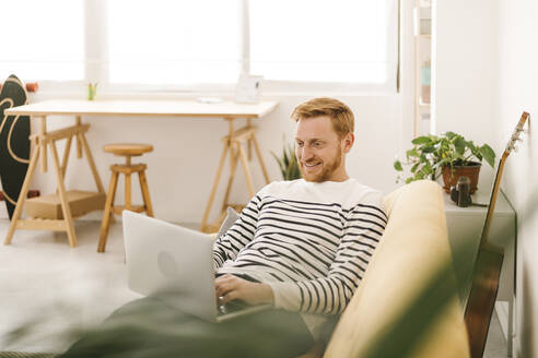 Glücklicher junger Mann mit Laptop auf dem Sofa zu Hause - XLGF02896