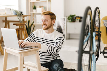 Happy man typing on laptop by bicycle at home - XLGF02901