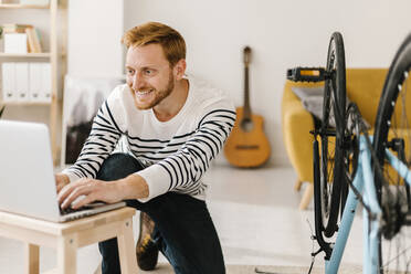 Smiling young man using laptop by bicycle in living room at home - XLGF02902