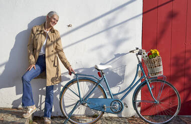 Lächelnde Frau mit Fahrrad vor einer Mauer stehend an einem sonnigen Tag - VEGF05497