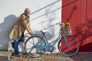 Frau stützt sich auf ein Fahrrad und steht vor einer Mauer an einem sonnigen Tag - VEGF05498