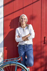 Smiling woman with arms crossed leaning on red door - VEGF05502