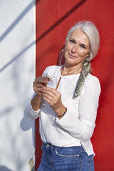 Smiling woman holding flower standing in front of red door on sunny day - VEGF05504