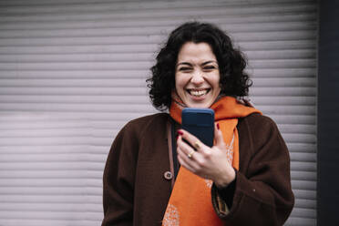 Happy young woman with mobile phone standing in front of corrugated shutter - AMWF00205