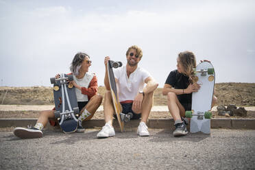 Happy friends with skateboards sitting at roadside on sunny day - FBAF01849