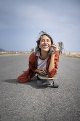 Glückliche junge schöne Frau liegt über Skateboard auf der Straße - FBAF01851