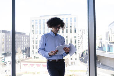 Businessman reading document leaning on glass wall in office - ASGF02200