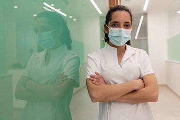 Nurse wearing protective face mask standing with arms crossed by glass wall in corridor of hospital - DLTSF02870