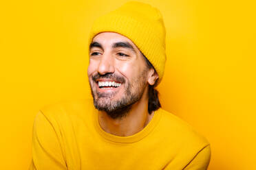 Glücklicher bärtiger Mann mit dunklem Haar, der wegschaut, während er auf gelbem Hintergrund in einem hellen Studio steht - ADSF33970