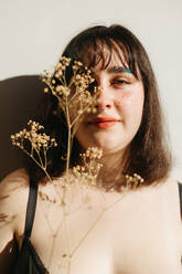 Positive plus size female in lingerie standing looking at camera on white background with dried twig in hand in room with bright sunlight - ADSF33973