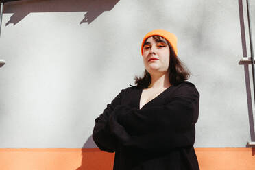 From below young female with green dyed hair and wearing a beanie in black jacket looking at camera while standing with arms crossed near orange and grey wall on sunny street - ADSF33976