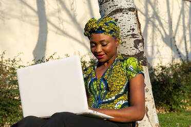 Afrikanische Freiberuflerin lächelt, während sie an einem sonnigen Tag im Park in der Nähe eines Baumes sitzt und auf ihrem Netbook surft - ADSF34014