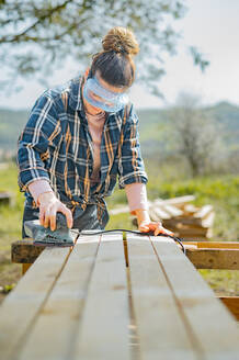 Konzentrierte Handwerkerin mit Schutzbrille bei der Arbeit mit einem speziellen elektrischen Schwingschleifer zum Glätten von Holzbrettern an einem Sommertag auf dem Land - ADSF34018