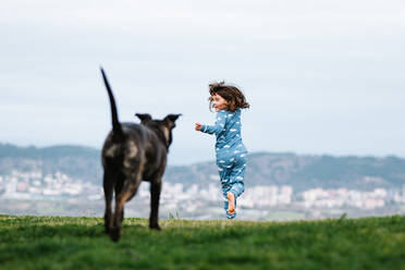 Full body back view of unrecognizable girl in pajama playing with American Pit Bull Terrier dog on grassy lawn against hills with residential buildings - ADSF34020