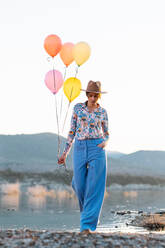 Nachdenkliche Frau mit Hut, die ein Bündel bunter Luftballons hält, während sie am Ufer eines plätschernden Sees steht und nach unten schaut - ADSF34023