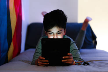 Arabischer Junge liegt auf dem Bett und surft im Internet auf einem Tablet in einem dunklen Raum, der mit einer Regenbogenflagge geschmückt ist - ADSF34033