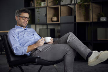 Geschäftsmann hält Kaffeetasse sitzt auf Stuhl im Büro - RBF08631