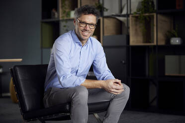 Happy businessman wearing eyeglasses sitting on chair in office - RBF08634