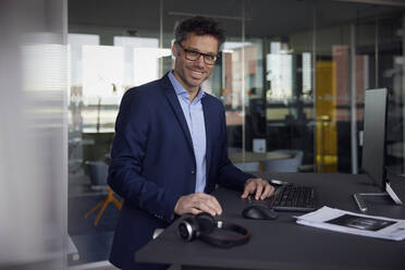 Smiling businessman wearing eyeglasses standing at desk in office - RBF08642