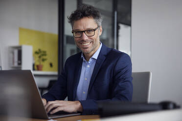 Glücklicher Geschäftsmann sitzt mit Laptop am Schreibtisch im Büro - RBF08648