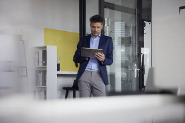 Businessman using tablet PC leaning on glass door at work place - RBF08655
