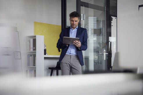 Geschäftsmann, der einen Tablet-PC benutzt und sich an eine Glastür am Arbeitsplatz lehnt - RBF08655