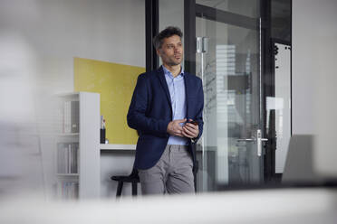 Thoughtful businessman leaning on glass door in office - RBF08656