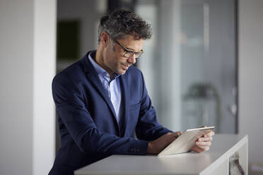 Businessman wearing eyeglasses using tablet PC standing at desk in office - RBF08660