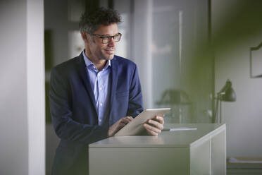 Thoughtful businessman with tablet PC standing at desk in office - RBF08661