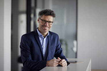 Happy businessman wearing eyeglasses standing at desk in office - RBF08662