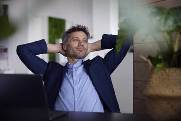 Smiling thoughtful businessman with hands behind head at work place - RBF08667