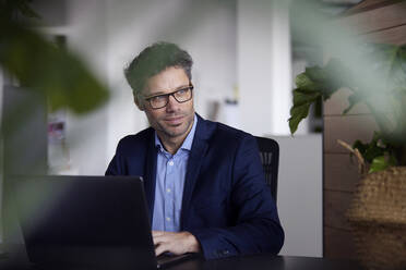 Geschäftsmann mit Brille sitzt mit Laptop am Schreibtisch im Büro - RBF08672