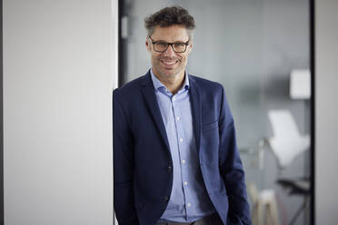 Smiling businessman wearing eyeglasses leaning on wall at work place - RBF08676