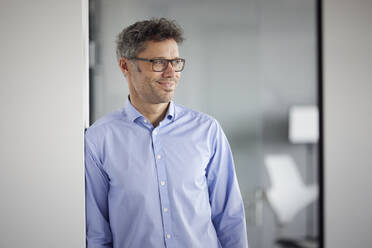 Smiling businessman wearing eyeglasses standing by wall at work place - RBF08678