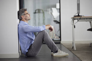 Businessman with eyes closed sitting by wall in office - RBF08679