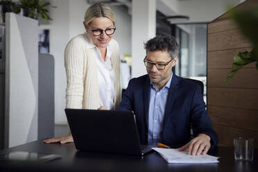 Businessman using laptop sitting by colleague standing at desk in office - RBF08687